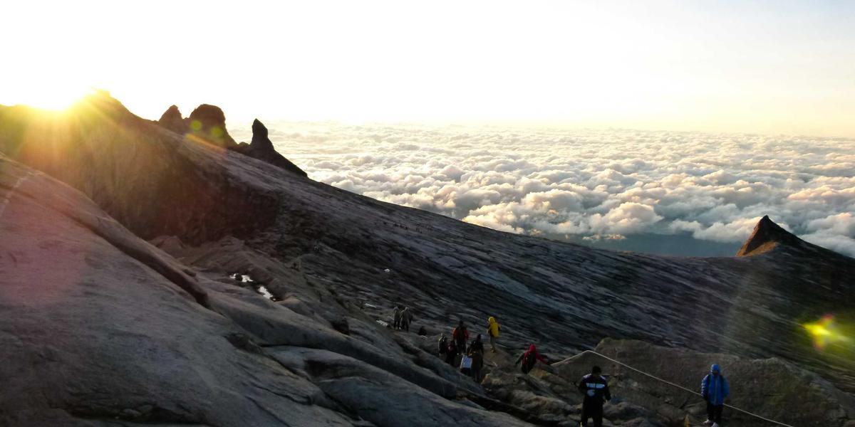 Hikers climbing a rocky mountain ridge at sunrise, above a sea of clouds.