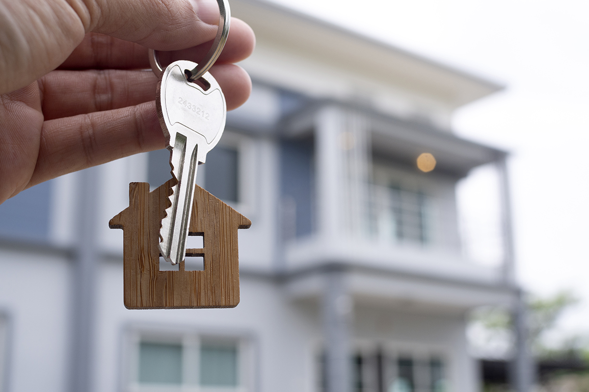 Key on a ring held up with a wooden house-shaped tag, in front of a modern home (symbolizing home ownership).