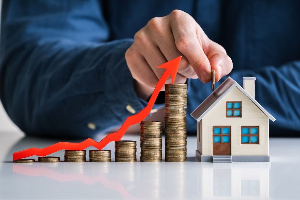 Hands stack coins beside a small house model, with a red upward arrow signaling rising real estate investment.