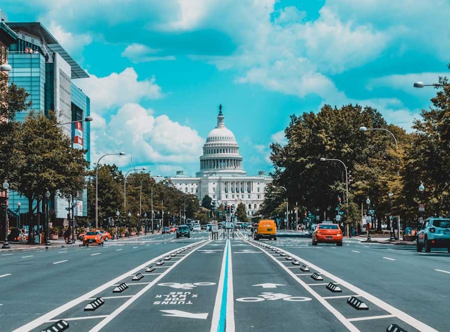 Capitol building centered in the distance with a street and bike lane in the foreground, trees lining the road, and traffic on a sunny day.