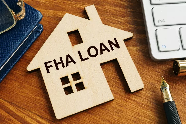 Wooden house cutout labeled 'FHA LOAN' on a wooden desk beside a keyboard and a fountain pen.