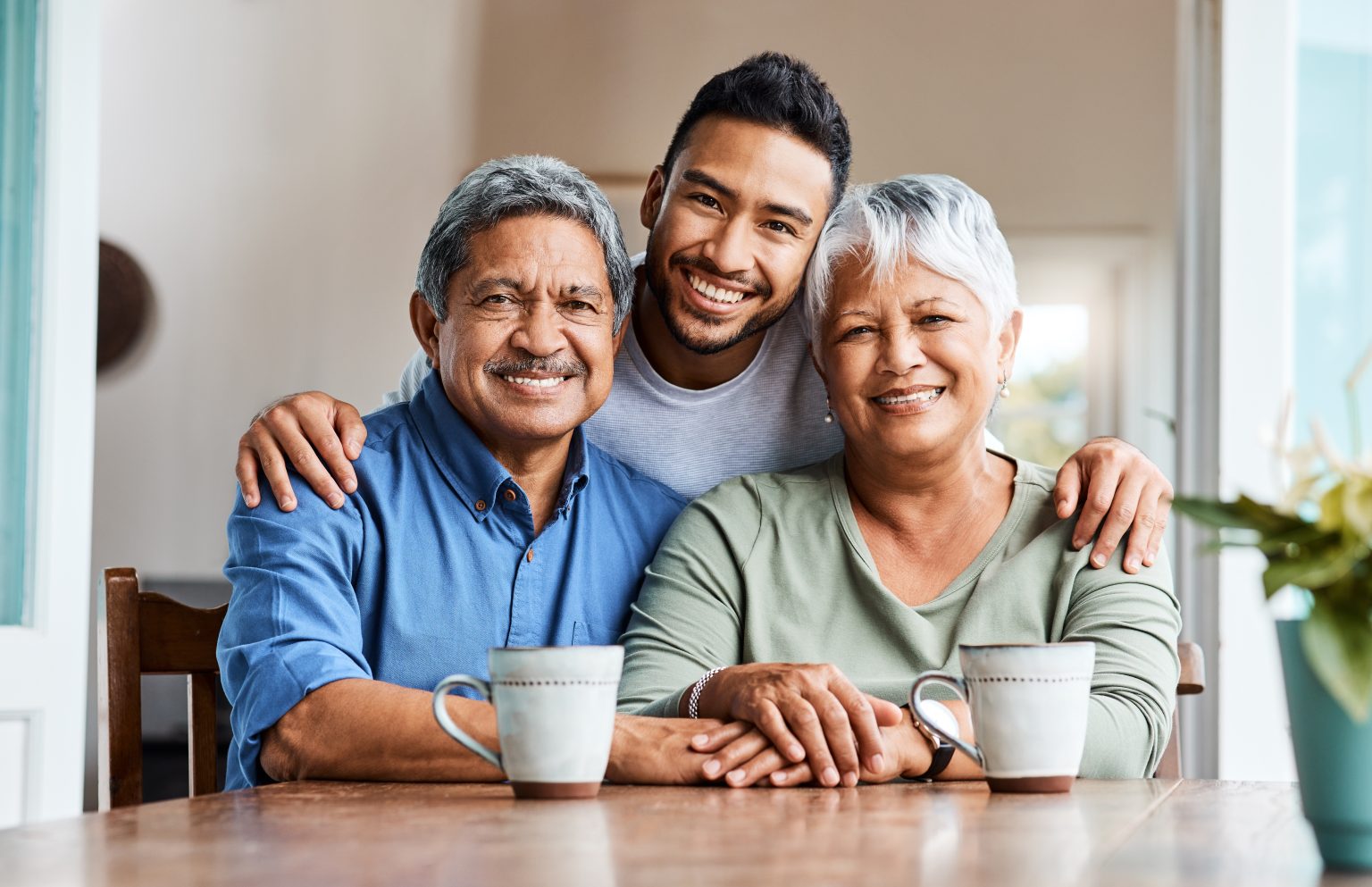 Grandfather, grandmother, and their adult son smiling at the camera, arms around each other, seated at a table with two mugs in front.